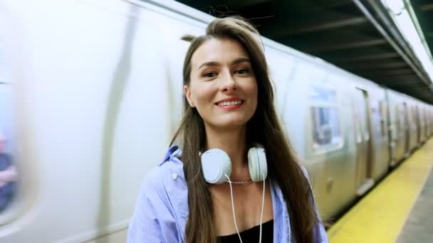 カメラの笑顔を見ながら地下鉄駅に立っている美しいかわいいコーカサス女の子の肖像画 バックグラウンドで電車でヘッドフォンを持つ魅力的な魅力的な若い女性 地下コンセプト — ストック動画