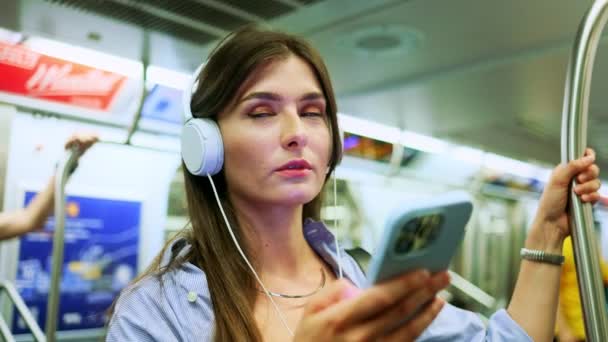 ヘッドフォンで音楽を聴き 地下鉄の自動車旅行でスマートフォンを保持する若い女の子は 無線インターネット接続を使用しています 携帯電話でオンラインチャット メッセンジャー 地下鉄でビデオを見る — ストック動画