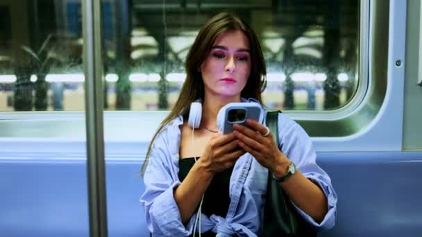 穿着无线耳机的年轻女人坐在火车上 带着智能手机 看着手机屏幕 漂亮的女孩通过手机在网上搜索信息 安静宁静的地铁 — 图库视频影像