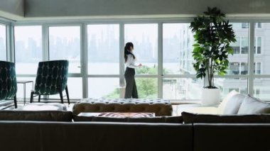 Gökdelen binasından panoramik manzara. Yeni modern ofisin penceresinin yanında duran profesyonel iş kadını akıllı telefonla konuşuyor. İletişim, yönetim ve üretken çalışma kavramı.