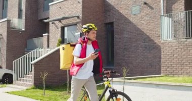 Bisikletiyle müşteriye yürürken akıllı telefon kullanan koruyucu kasklı genç bir işçi. Termal sırt çantası takan yakışıklı bir erkek kurye özel uygulamadan müşteri adresi arıyor..