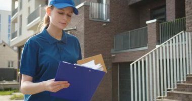 Beyaz, genç, güzel bir kadın. Şapkalı bir teslimatçı. Elinde karton kutuyla sokakta yürüyor ve belgelere bakıyor. Paket konsepti olan güzel bir kurye.