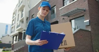 Kurye caddede yürüyor ve dosyaya dökümanlarla bakıyor. Karton kutuyu elinde taşıyan teslimatçı kadın. Kartpostal teslimatı. Teslimat konsepti