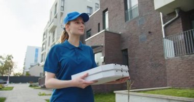 Beyaz, güzel bir kadın, şapkalı bir teslimatçı sokakta yürüyor ve pizza kutusuyla karton kutu taşıyor. Açık hava paketli güzel bir kurye.