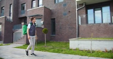 Büyük termal sırt çantalı genç bir erkek kuryenin caddede yürüyüp müşteri adresini ararken yan görüntüsü. Kafkasyalı kurye adres arıyor. Çalışma konsepti