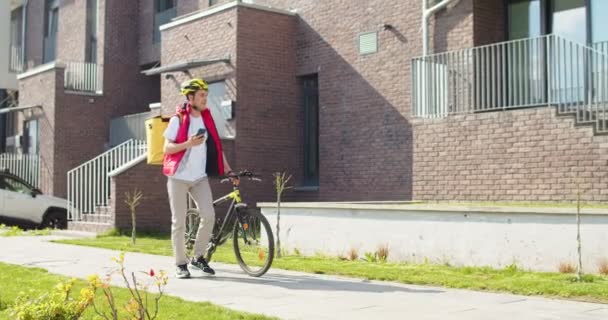 お客様に食べ物を届けるサーマルバックパック付きの魅力的な若い男性宅配便 自転車で歩く安全ヘルメットを身に着けている白人配達員 商品配送コンセプト — ストック動画