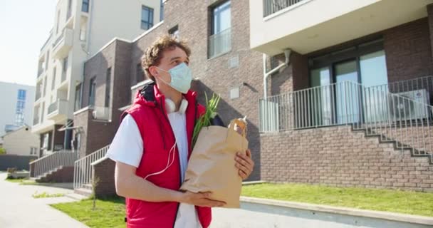 白人年轻人 戴着医疗面罩的送货工人穿过街道 提着装有新鲜食物的包裹 男性快递员将提供食品杂货概念 — 图库视频影像