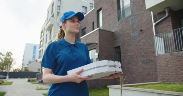 白人の若い美しい女性 キャップの配達労働者は通りを歩き ピザでカートン箱を運びます 女性の美しい宅配便小包屋の屋外 — ストック動画