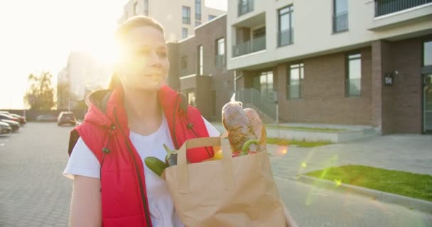 白人女孩 送货员穿过街道 提着装有新鲜食物的包裹 美丽的女快递员将在夏夜运送食品 — 图库视频影像
