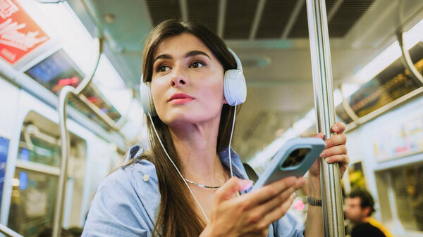 Smiling charming woman wearing wireless headphones while moving in tram. Big city lifestyle, transportation. Female chatting online in mobile phone messenger watch video in metro.