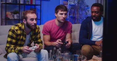 Yakışıklı, mutlu, Afro-Amerikan ve beyaz erkekler video oyunları oynayıp, kanepede oturup gülüşerek eğleniyorlar. Boş zaman konsepti. Oyuncuların en iyi arkadaşları geceleri konsolda video oyunu oynarlar