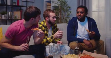 Neşeli gülümseyen beyaz ve Afrikalı Amerikalı erkek arkadaşlar pizza yiyor ve TV karşısında futbol maçı izleyip eğleniyorlar. Erkekler geceleri kanepede oturup futbol maçı izler.