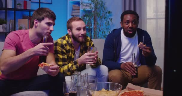 三位年轻快乐的混血种族男性朋友晚上在家里聚在一起 一边吃披萨 一边喝酒 一边聊天 玩得很开心 男人们坐在沙发上看体育频道 — 图库视频影像