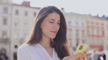 Neşeli, heyecanlı, günlük kıyafetler giyen beyaz kız gezgin gülümseyerek ve sokakta akıllı telefon sosyal medya uygulamaları kullanarak. Sinema merkezi konseptinde seyahat eden kadın.