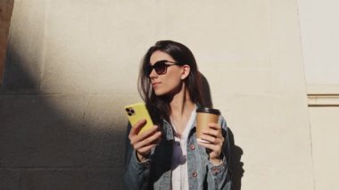 Genç gülümseyen esmer kız sokakta duvarın yanında oturup mesaj gönderirken günlük kıyafetler giyip akıllı telefon kullanıyor. İnsanlar ve teknoloji kavramı