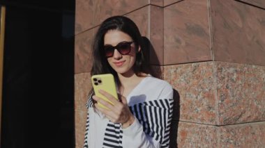 Genç gülümseyen esmer kız sokakta duvarın yanında oturup mesaj gönderirken günlük kıyafetler giyip akıllı telefon kullanıyor. İnsanlar ve teknoloji kavramı