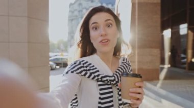 Selfie kamera görüntüsü. Heyecanlı kız kameraya bir şey söylüyor ve yazın sokakta yürürken elinde kahve tutarken duygusal bir şeyler hissediyor.