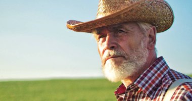 Güneşli bir günde mavi gökyüzü arka planındaki tarlaya bakan sakallı yaşlı adamın portresini yakından çek. Yaz doğasındaki yaşlı çiftçinin yüzü. Gerçek insan çiftçiliği kavramı