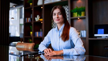 Başarılı ve profesyonel genç bir kadının portresi ünlü bir şirkette masada oturmuş resmi kıyafetler giymiş kameraya bakıp gülümsüyor. Zarif çekici iş kadını. İşveren kavramı.