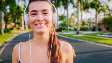 Formda kız portresi idman kıyafetleri içinde koşmaya hazırlanıyor kameraya gülümsüyor. Güzel, mutlu sporcu kadın kulaklıkla müzik dinlemekten zevk alıyor. Müzik konsepti.