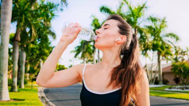 Kadın içme suyu içiyor ve kameraya bakıyor. Sabah koşusu. Palmiye ağaçlarının arasında koşan esmer ve çekici bir kız. Su şişesiyle koşan genç ve güzel bir kadın. Spor koşusu konsepti.