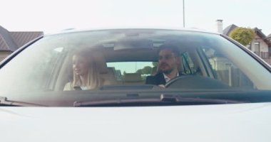 Sürücü koltuğunda oturan beyaz bir adam yola çıkıp onun güzergahını düşünürken pencereye bakıyor. Karısı yakınlarda oturuyor.