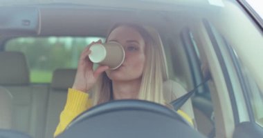 Genç, mutlu, beyaz bir kadın yolda çay ya da kahve içerek araba sürüyor. Müzik dinlediğim için mutluyum. Keyfin yerinde. Afiyet olsun. Arabaların içi. Ön manzara. Kapat..