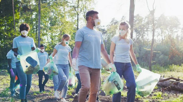 Multiracial, diverse group of volunteers collecting garbage, waste. Active environmentalists wearing protective masks walking together chatting with each other. Volunteering, global concern concept