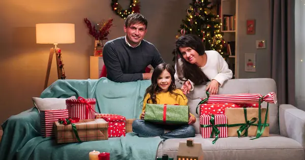 Happy caring Caucasian parents mom and dad giving xmas wrapped gift to little joyful cute surprised girl who received many presents on New Year. Holiday tradition. Merry Christmas concept