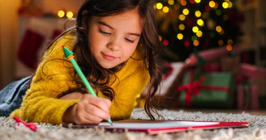 Kafkas neşe dolu küçük kızın yakın çekimleri, dekore edilmiş bir odada, arka planda parıltılı ışıklar olan kağıtların üzerine uzandı. Çocuk Noel Baba 'ya mektup yazıyor. Noel tatili konsepti