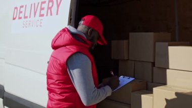 Afro-Amerikalı teslimatçı. Paket kutusunu alıcıya götürüyor. Kurye kutuları işaretlemiş, teslimat aracının yan kapısını kapatmış ve online siparişleri teslim etmek için gitmiş..
