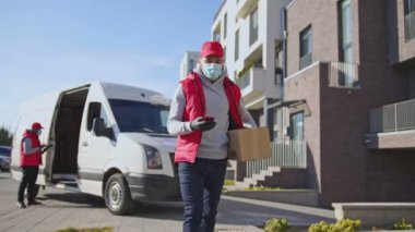 Yüz maskeli, eldivenli, paket kutusunu taşıyan, sokakta telefonla dolaşan beyaz bir teslimatçı. Bir müşteriye posta paketi teslimatı için kurye yolda..
