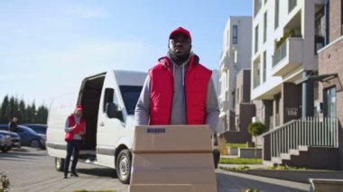 Afro-amerikan kurye bir kamyonetten bir yük arabasında paket kutuları taşıyor. Bir müşteriye posta paketi teslimatı için kurye yolda..