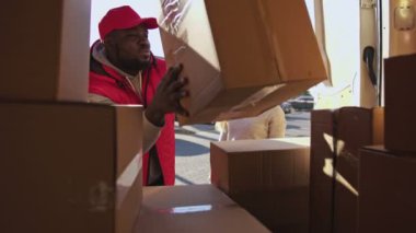 Kırmızı üniformalı Afro-Amerikan kurye bir minibüsten paket kutusunu alıp bir müşteriye veriyor. Beyaz bir kadın sipariş aldı ve bir tablet imzaladı..