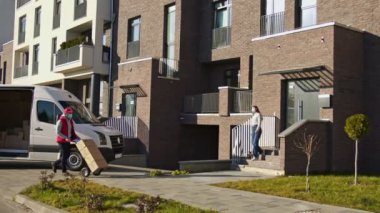 Yüz maskeli beyaz kurye bir kamyonetten bir paket kutusunu taşıyıp bir müşteriye veriyor. Kadın evinin yakınında bir sipariş alıp bir tablet klasörü imzaladı..