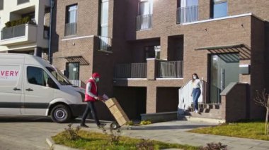 Yüz maskeli beyaz kurye bir kamyonetten bir paket kutusunu taşıyıp bir müşteriye veriyor. Kadın evinin yakınında bir sipariş alıp bir tablet klasörü imzaladı..