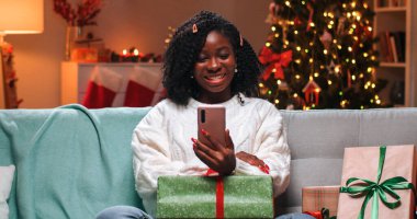 Noel akşamı dekore edilmiş odada dinlenen mutlu Afrika kökenli güzel bir kadının portresi. Yanında cep telefonuyla parlayan ağaçtan görüntülü sohbet hediyeleri. Yeni yıl konsepti