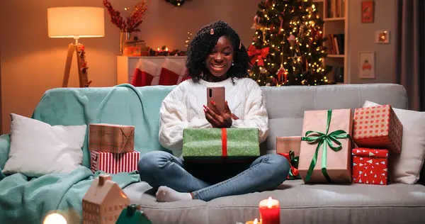 Cheerful beautiful African American female resting in decorated room on Christmas evening with xmas gifts with glowing tree waving hand and speaking on video call on cellphone. New Year concept