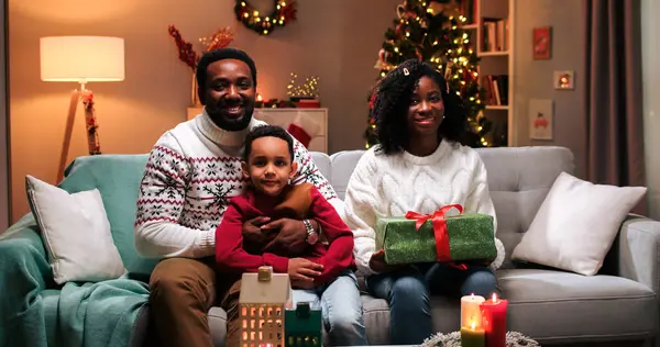 Portrait of happy African American family couple with cute small kid son sitting in decorated room at home with xmas gift, smiling to camera on Christmas. New Years eve celebration concept