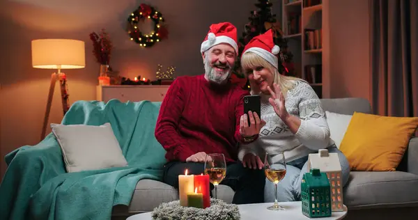 Joyful Caucasian family man and woman drinking wine and clinking glasses while speaking on video call with kids on smartphone celebrating New Year while sitting in decorated room in evening. Xmas mood