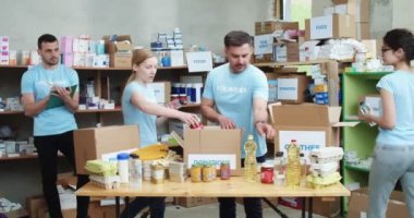 Mavi gönüllü tişörtlü üç aktif genç depoda bağış kutularını topluyor. Erkek lider arkada duruyor ve not alıyor..