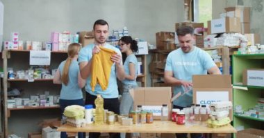 Mavi üniformalı iki beyaz adam depoda bağış kutularını toplarken iki kadın da raflarda yiyecek teftişi yapıyor. Hayır kurumu için çalışan gençler.