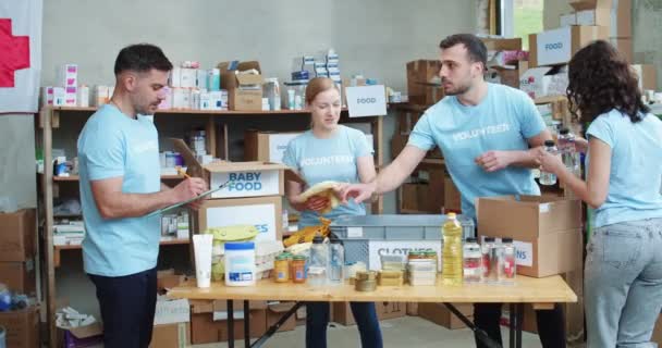 一队身穿蓝色志愿T恤衫的年轻人在食品库仓库里整理和包装捐赠的物品 站在剪贴板上做广告的白人男性策展人 — 图库视频影像