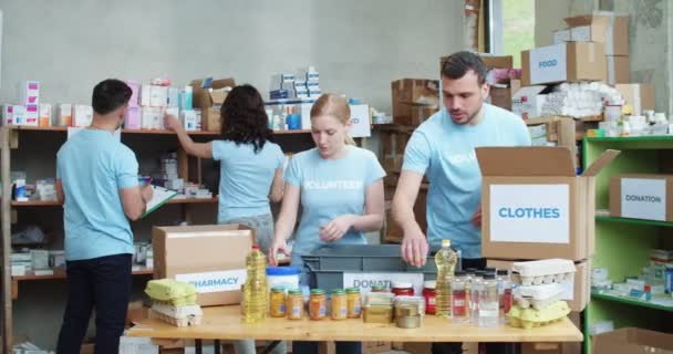 身穿蓝色T恤衫的白人男女积极分子在慈善组织的仓库里一起工作 自愿人员小组在架子上整理供应品 在纸板箱中包装 — 图库视频影像