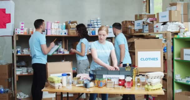 身穿蓝色T恤的热诚的高加索志愿者一起站在仓库和包装捐赠箱 合作帮助有需要的人的青年积极分子团体 — 图库视频影像