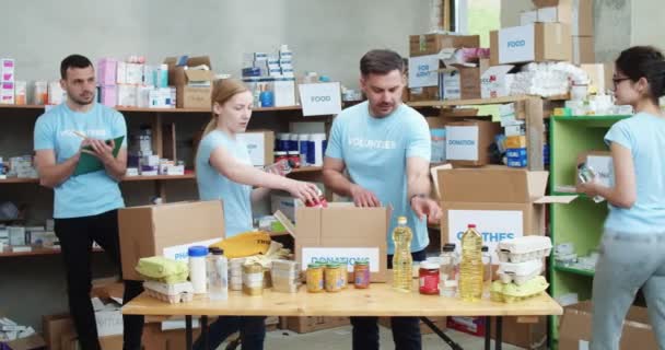 Три Активных Молодых Человека Синих Волонтерских Футболках Упаковывают Коробки Пожертвований — стоковое видео