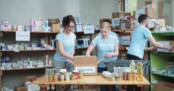 一群年轻的志愿者为慈善中心整理捐赠的食品和罐头食品 男性领导协调团队工作时 在仓库里走来走去 并在剪贴板上做笔记 — 图库视频影像