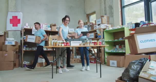 デジタルタブレットで働く青いシャツの女性労働者は フードバンクの倉庫でプロビジョニングボックスを調べている 赤十字のためのチャリティファンドで一緒に働くボランティアのグループ — ストック動画