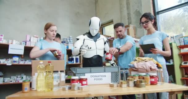 必要としている人々に箱を準備するために人工ロボットと協力する食品銀行労働者のチーム ヒューマノイドパッキングボックスの間 タブレットやソートグッズを使用する若いボランティア — ストック動画