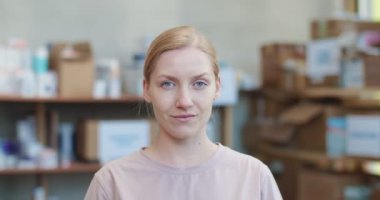 Kendine güvenen, beyaz, sarı saçlı bir kadın bağış malzemeleriyle rafların arasında duruyor ve kameraya bakıyor. İnsanlar, hayır kurumu ve gıda bankası kavramı.